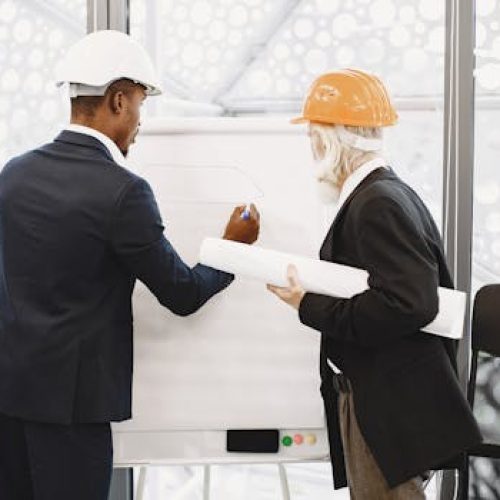 Two architects in hard hats working on a blueprint indoors, highlighting teamwork.
