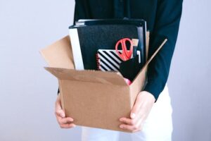 Woman carrying cardboard box with office items like scissors and notebooks.