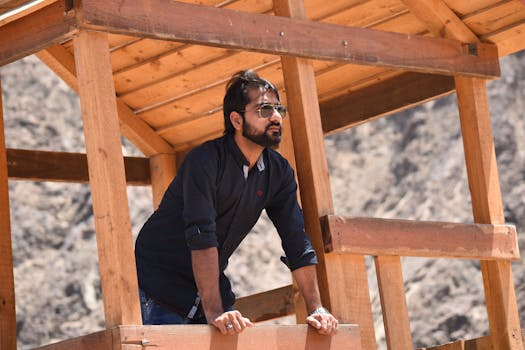 A man with sunglasses leans on a wooden structure outdoors, under clear daylight.